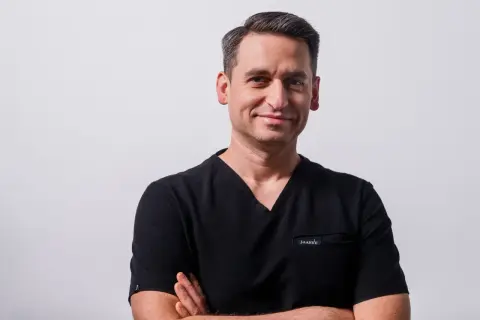 Mężczyzna w czarnym scrubie, ze skrzyżowanymi rękami, uśmiecha się do kamery.
