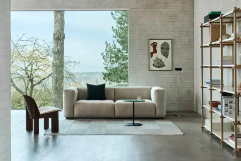 Gemütliches Wohnzimmer mit einem Sofa in millennial grey, einem Holzsessel und einem Bücherregal vor einem Fenster mit Blick ins Grüne.