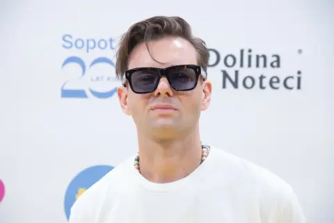 Rubens piosenkarz w okularach przeciwsłonecznych i naszyjniku z koralików, pozuje na tle napisu "Sopot 20 Lat" i "Dolina Noteci".