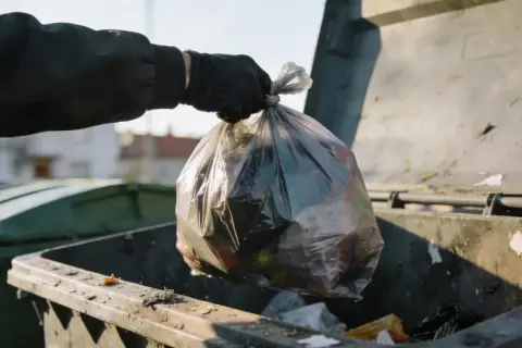 Ręka w czarnej rękawiczce wrzuca worek ze śmieciami do kontenera. Zastanawiamy się, ile zapłacimy za wywóz śmieci.