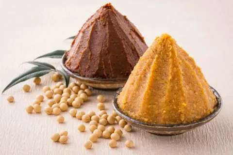 Due ciotole con miso, uno scuro e uno chiaro, circondate da fagioli di soia. Il miso è una pasta fermentata giapponese.