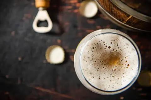 Un bicchiere di birra con una schiuma dorata e bollicine, accanto a un apribottiglie e tappi su uno sfondo scuro. La birra senza schiuma è un piacere per gli occhi.