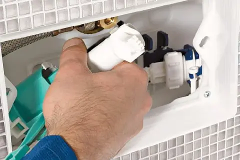 Dłoń montuje mechanizm spłuczki podtynkowej w białej obudowie. Widoczne elementy hydrauliczne i zielony zbiornik.