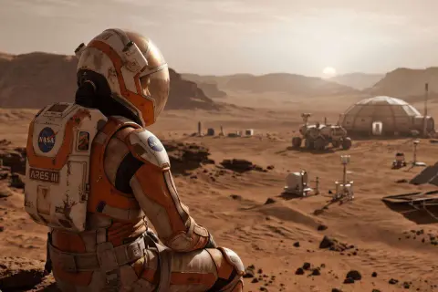 Astronaut w pomarańczowym skafandrze NASA z napisem ARES III siedzi na Marsie, patrząc na bazę i łazik. Scena jak z filmów z Mattem Damonem.