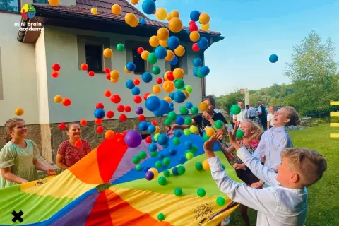 Radosne zabawy dla dzieci na urodziny: kolorowe piłeczki latają w powietrzu nad tęczowym spadochronem, a dzieci śmieją się z radości.