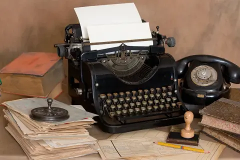 Escritorio antiguo con máquina de escribir, teléfono negro, libros y papeles. Un lápiz amarillo descansa sobre documentos, evocando la creación de dibujos con máquina de escribir.