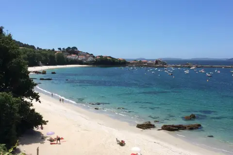La **playa de Aguete** luce un día soleado con aguas cristalinas y barcos fondeados. Gente disfruta de la arena blanca y el mar azul.
