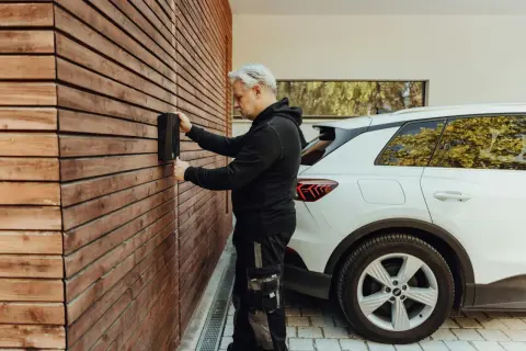 Un homme installe une borne de recharge pour voiture électrique sur un mur en bois. La question "peut on installer soi meme une borne de recharge" se pose.