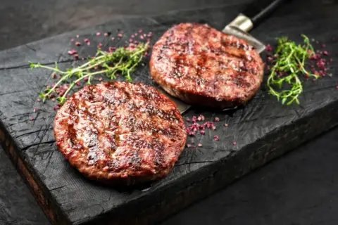 Dwa soczyste, grillowane kotlety na burgery na drewnianej desce z ziołami i różowym pieprzem.