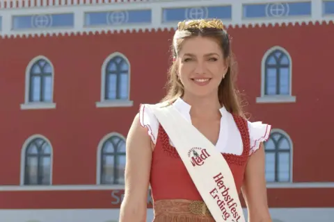 Eine strahlende junge Frau mit Krone und Schärpe "Herbstfest Erding" vor einem roten Gebäude.