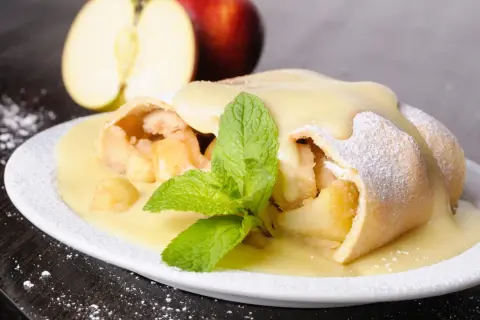 Ein köstlicher Apfelstrudel mit Eiermilch, bestäubt mit Puderzucker und garniert mit Minze. Daneben liegen Äpfel.