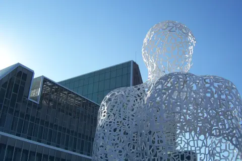 Biała, ażurowa rzeźba postaci z liter, przypominająca monumentalizm, na tle nowoczesnych budynków i błękitnego nieba.