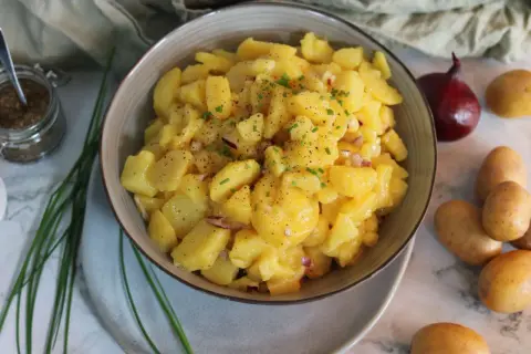 Schüssel mit leckerem Kartoffelsalat ohne Mayo, garniert mit Schnittlauch und Pfeffer. Daneben liegen frische Kartoffeln und eine rote Zwiebel.