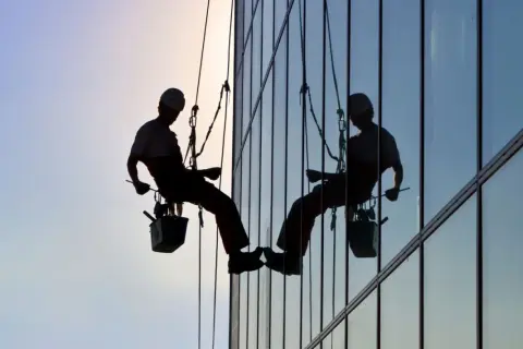 Siluetas de operarios realizando limpieza de cristales en edificios y locales, suspendidos por cuerdas.