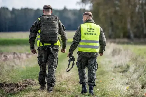Dwóch żołnierzy Wojsk Obrony Terytorialnej w kamizelkach odblaskowych idzie przez pole. Rezygnacja z WOT może mieć poważne konsekwencje.