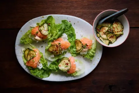 Läckra asiatiska tapas med lax, ris och gurka i salladsblad. En skål med marinerad gurka står bredvid.
