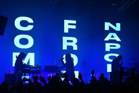 Sul palco, silhouette di musicisti con tastiere e microfoni, mentre sullo sfondo campeggia la scritta "COM FROM NAPOLI" in lettere luminose.