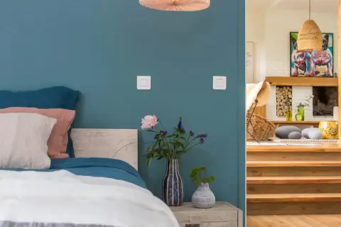 Un lit douillet avec des coussins roses et blancs sur un mur couleur bleu canard. À côté, un escalier en bois mène à un salon cosy.
