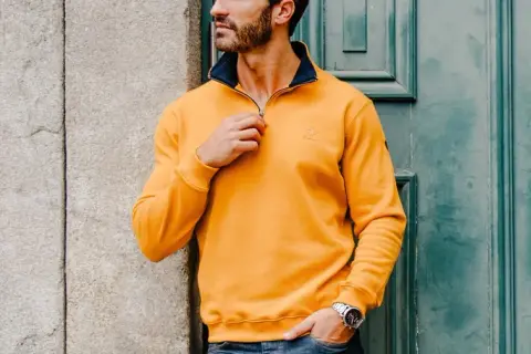 Un homme ethnic blue porte un pull jaune vif avec un col bleu marine, ajustant la fermeture éclair. Il est appuyé contre un mur en pierre.