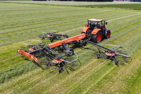 Pomarańczowy traktor zgrabnie manewruje po polu, ciągnąc szeroką zgrabiarkę do traktorka, która sprawnie formuje pokosy zielonki.