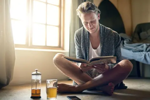 Hombre joven leyendo un libro, disfrutando del **slow living** junto a una ventana con luz natural y una taza de té.