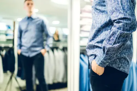 Mann im Spiegel prüft seinen lässigen kleidungsstil herren. Blaues Hemd mit Muster, schwarze Hose.
