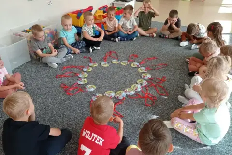 Dzieci siedzą w kółku na dywanie, patrząc na okrąg z zawieszkami. Może to być część zabawy, jak w "Mam chusteczkę haftowaną".