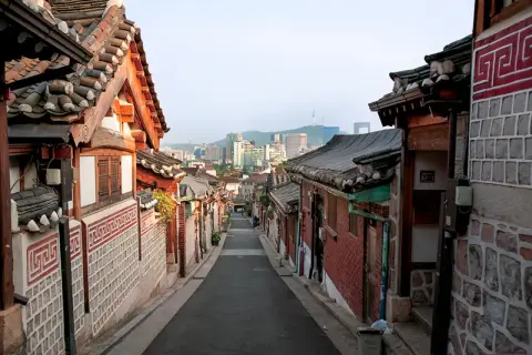 En smal gata kantad av traditionella hus med tegeltak leder mot en modern stadssilhuett i Seoul, Sydkorea.