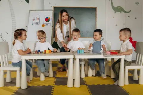 Nauczycielka bez wykształcenia pedagogicznego prowadzi zajęcia z dziećmi w przedszkolu. Dzieci siedzą przy stolikach, bawiąc się klockami i kartami.