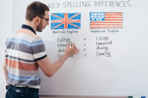 Profesor explica las diferencias de ortografía entre los tipos de inglés: británico y americano, con ejemplos escritos en la pizarra.