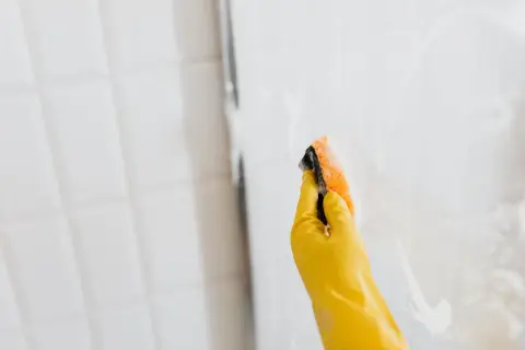 Mano con guante amarillo limpiando mampara de ducha con esponja.