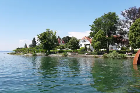 Friedrichshafen am Bodensee: sonnige Uferpromenade mit weißen Villen, grünen Bäumen und einer rostigen Skulptur im Wasser.