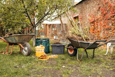 Gartenarbeit im Herbst: Schubkarren voller Laub und Äste warten darauf, kompost umsetzen zu helfen. Ein Eimer mit Küchenabfällen steht bereit.