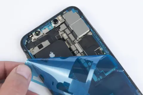 Dłoń odkleja niebieską folię od wnętrza iPhone'a XR z widoczną baterią.
