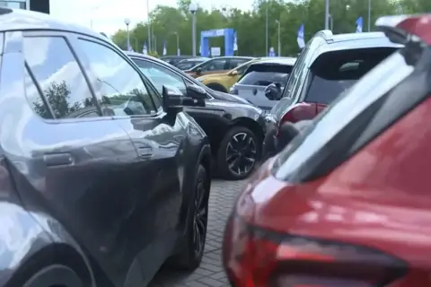 Rzędy samochodów, w tym czerwony i szary SUV, czekają na swoich właścicieli. W tle widać bramę z logo, sugerującą salon samochodowy lub wydarzenie motoryzacyjne.