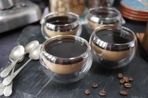 Fyra små glas med kaffe pannacotta, toppad med mörk sirap. Tre skedar ligger till vänster, och kaffebönor sprider sig på stenen.