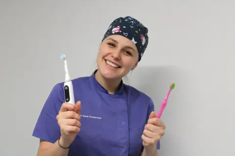 Sorriso smagliante di un dentista con cappellino a tema natalizio, che mostra uno spazzolino elettrico e uno manuale. Si può usare lo spazzolino elettrico con l'apparecchio.