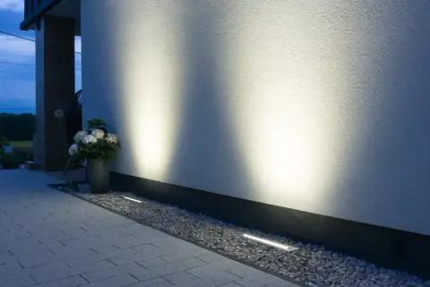 Eleganckie oprawy najazdowe subtelnie oświetlają ścieżkę i białą elewację domu, tworząc nastrojową atmosferę wieczorem.