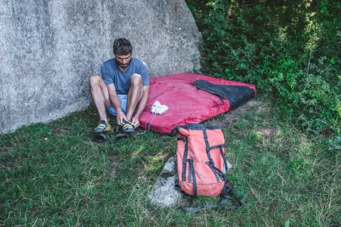 Man förbereder sig för kjugekull bouldering, spänner sina skor bredvid en tjock madrass och en orange ryggsäck.
