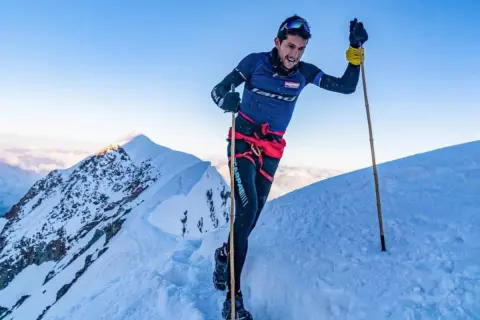 Manuel Merillas, le coureur de trail, franchit une crête enneigée, bâtons en main, sous un ciel bleu éclatant.