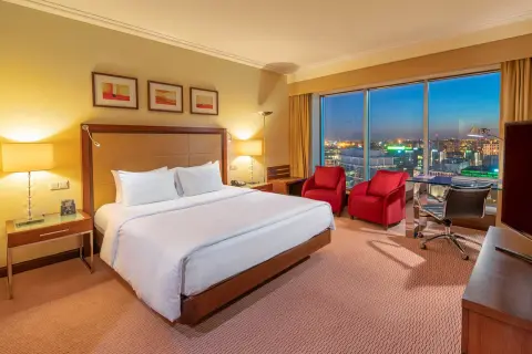 Przytulny pokój w hotelu Hilton Warszawa z widokiem na nocne miasto. Duże łóżko, dwa czerwone fotele i biurko.