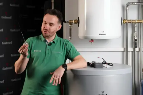 Mężczyzna w zielonej koszulce prezentuje element, który może być anodą tytanową, obok zasobnika wody.