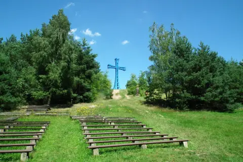 Niebieski krzyż na wzgórzu, najwyższy szczyt wielkopolski, otoczony drzewami. Poniżej amfiteatr z drewnianymi ławami.