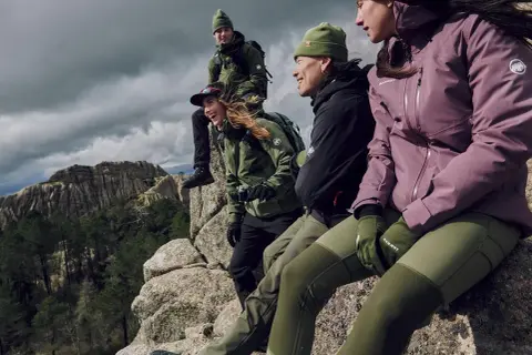 Grupa przyjaciół w zielonych i fioletowych kurtkach w góry, siedząca na skale, podziwiając krajobraz.