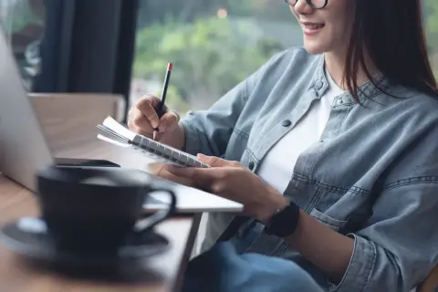 Kobieta pisze w notatniku, korzystając z prawa cytatu, by zanotować inspiracje. Obok kawa i laptop.