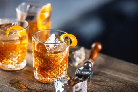 Tre glas med old fashioned drinkar, is och apelsinskal på träbord. En cocktailgaffel syns i bakgrunden.