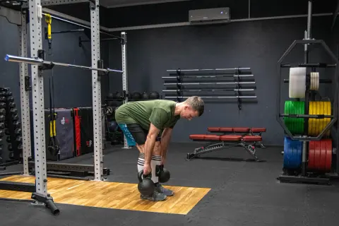 Mężczyzna wykonuje martwy ciąg rumuński z kettlebellami, ćwicząc mięśnie tylnej taśmy. W tle widać sprzęt do treningu siłowego, w tym sztangi i obciążenia.