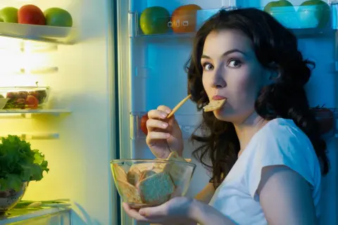 Une femme, prise par une **sensation de faim**, grignote des biscuits devant un réfrigérateur ouvert, rempli de fruits et légumes.