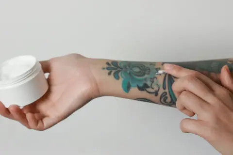 Pflege für dein Tattoo: Eine Hand hält eine Dose mit weißer Creme, während die andere Hand sie auf ein buntes Tattoo aufträgt. Kokosöl Tattoo-Pflege.