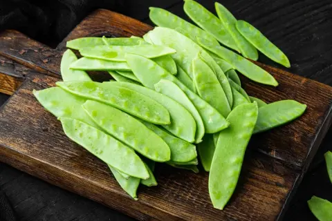 Un mucchio di taccole fresche, verdi e piatte, su un tagliere di legno scuro. Cosa sono? Deliziose!